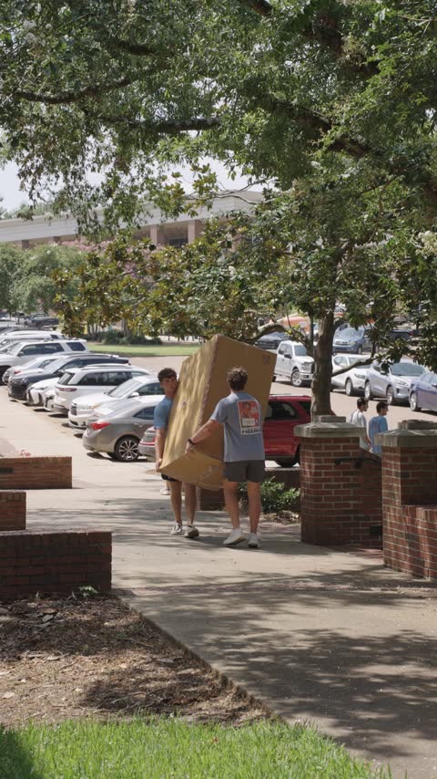 Two crew members carrying a large furniture box along the sidewalk past a row of parked cars in Oxford