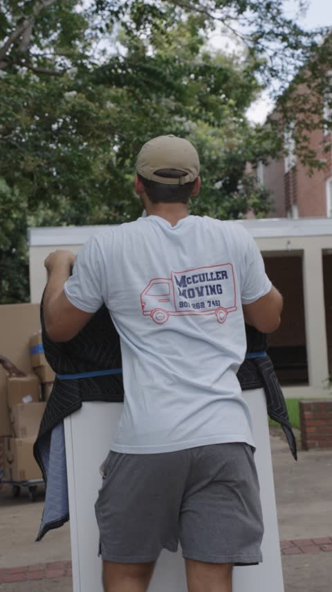 A crew member steering a wrapped wardrobe on a cart with a moving blanket over his shoulder