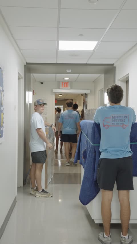 Crew members carrying a blanket-wrapped headboard through a residence hall corridor