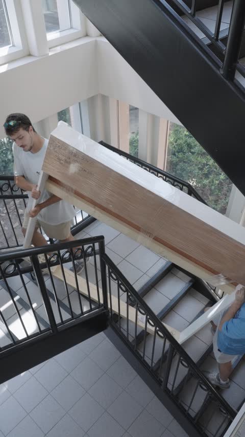 Two crew members carrying a shrink-wrapped wooden headboard down an apartment staircase