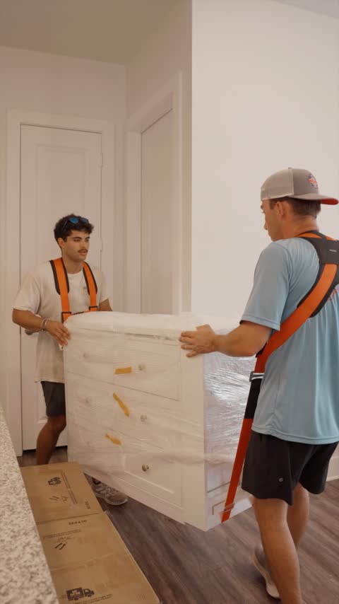 Two crew members lifting a shrink-wrapped white dresser with carrying straps