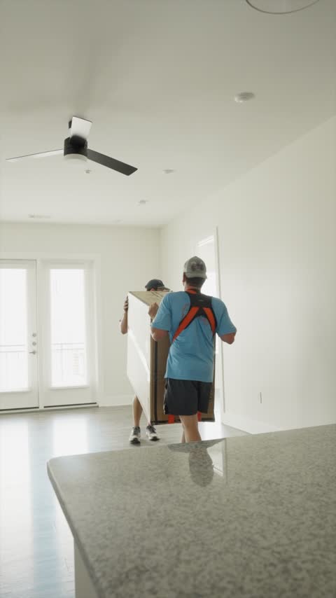 Two crew members carrying a flat-pack box into a bright, empty apartment