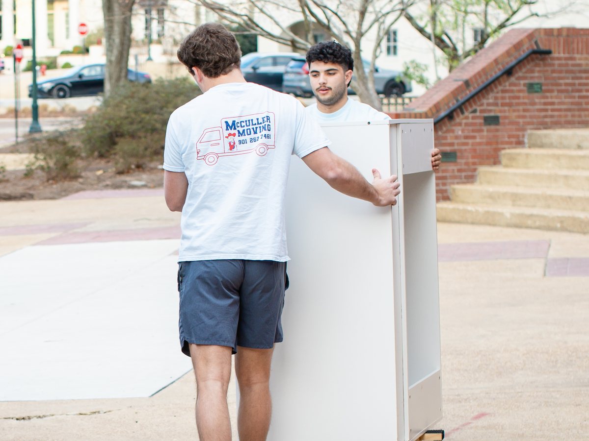 Two McCuller Moving crew members wheeling a white cabinet on a dolly across campus on move-out day
